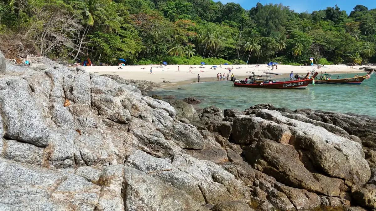 Longtail boats moored off a Phuket beach behind a rocky shoreline