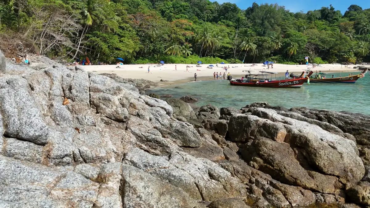 Longtail boats moored off a Phuket beach behind a rocky shoreline